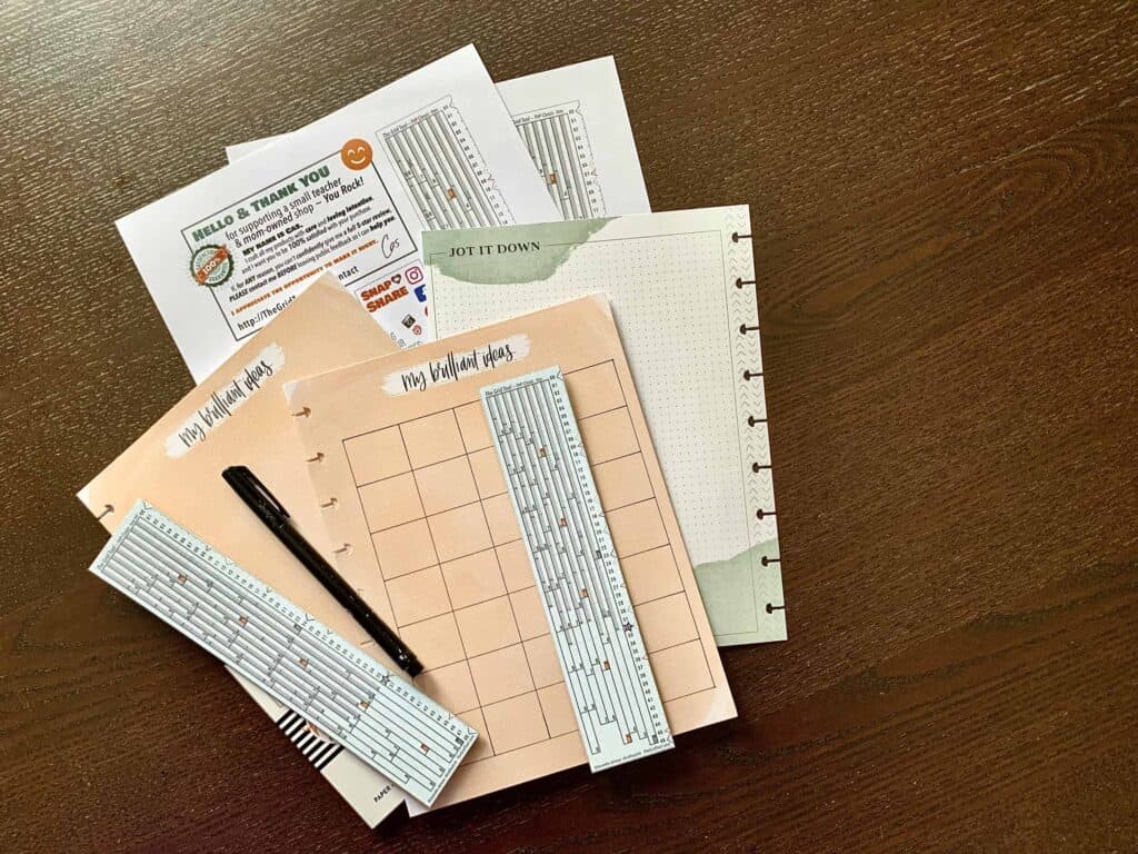 Photograph of several different styles of The Happy Planner Classic size Dot Grid paper with a 5x7 grid drawn on the top sheet and The Grid Tool in 2 sizes each laid over the top